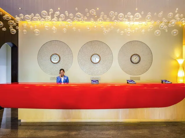 A receptionist in a blue jacket stands behind a red curved reception counter beneath a cluster of glass bubble lights, with three circular wall clocks behind her.