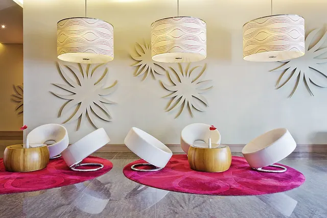A seating area with white swivel armchairs on pink rugs, wooden side tables with red flowers, sculpted wall decorations and three pendant lamps.