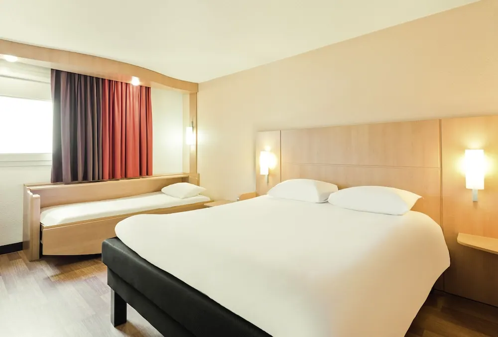 A bright hotel bedroom with a large double bed, wood-panel headboard, and a second single bed beneath the window.