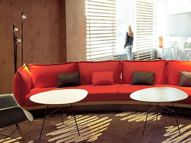 A bright hotel lounge has a long red sofa, round tables, armchairs and large windows with blinds.