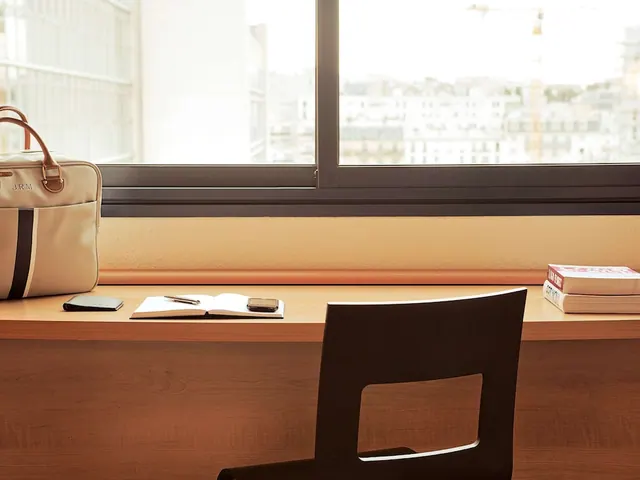 A desk with a chair, notebook, bag and books is positioned beside a large window overlooking city buildings.