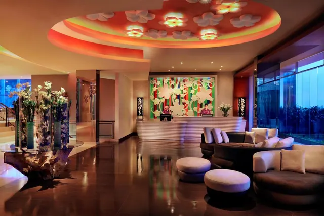 A hotel lobby with a reception desk behind a patterned feature wall, a flower-motif ceiling, lounge seating, and floral arrangements on a glass table.