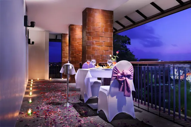 A dining table set for two with white chair covers and purple sashes stands on a terrace at dusk, with rose petals and candles on the floor and city lights visible beyond the railing.