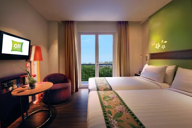 A hotel bedroom with two beds dressed in floral runners, a wall-mounted television, an armchair and a round side table, with glass doors opening onto a balcony with a view of trees.