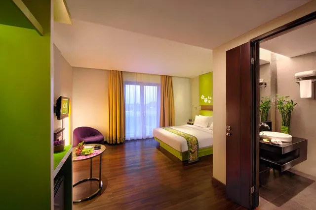 A hotel bedroom with a made double bed, a purple armchair, a round side table with fruit, a sliding door to the window, and a bathroom with a vessel sink visible through an open door.