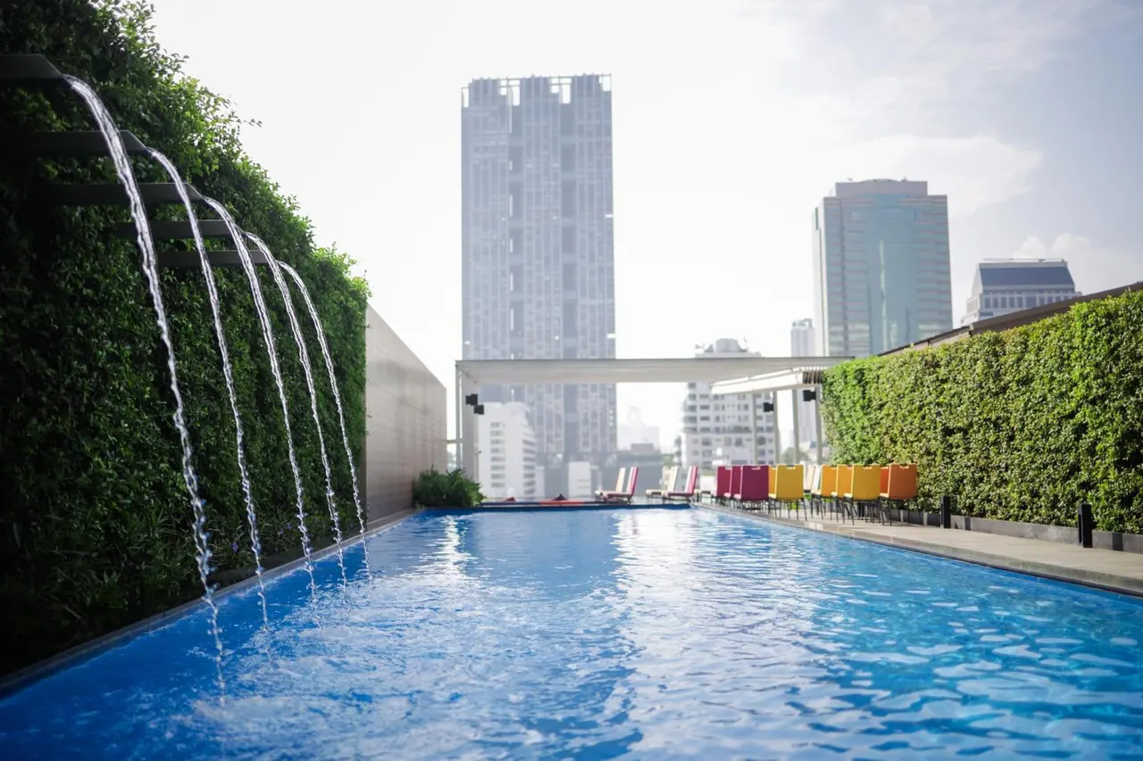 A long rooftop swimming pool with a water feature spilling from a green wall on the left, colourful lounge chairs on the right, and city towers in the background.