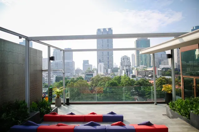 A rooftop terrace with red and blue cushioned seating, a glass railing, and a view over city towers and trees.