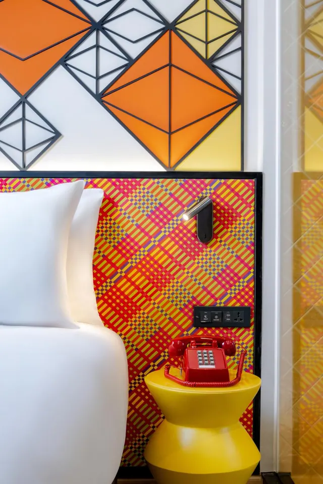 A bedroom corner shows a white bed beside a patterned orange and red headboard wall, with a yellow side table holding a red telephone.