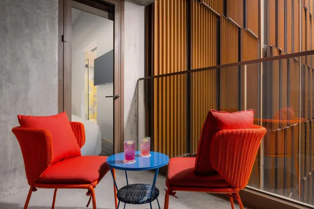 A small balcony with two orange wicker armchairs, a blue round table holding two drinks, and a glass door leading into the room.