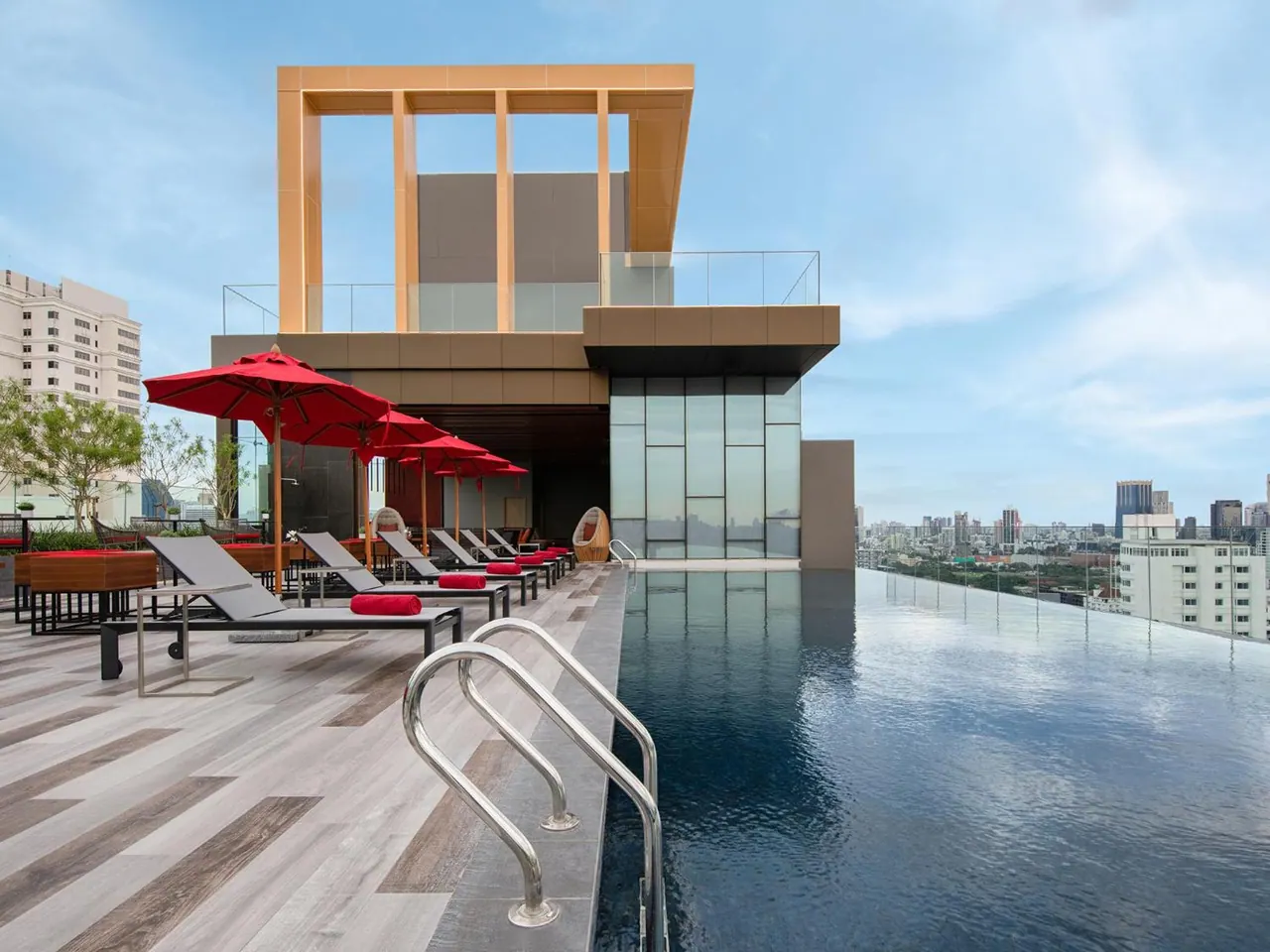 A rooftop infinity pool with red umbrellas and loungers overlooks a city skyline under a blue sky.