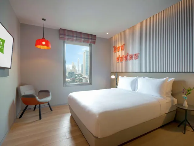 A bedroom with a made-up king bed, a grey armchair, a red pendant lamp, a wall-mounted television, and a window showing city skyscrapers.