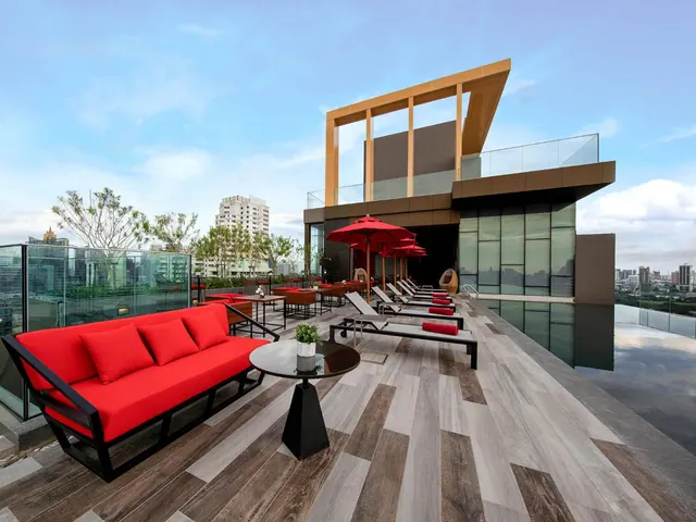 A rooftop terrace with red sofas, sun loungers, red parasols and a pool edge, with city buildings visible in the background.