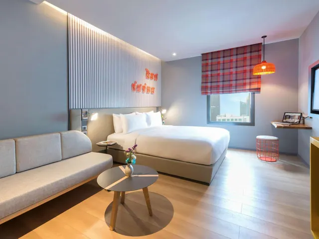 A hotel bedroom with a king bed, a grey sofa, a round coffee table, a wall-mounted desk and a window showing city buildings.