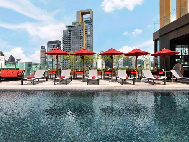 A rooftop pool with red umbrellas, grey sun loungers and a red sofa, with city towers in the background.