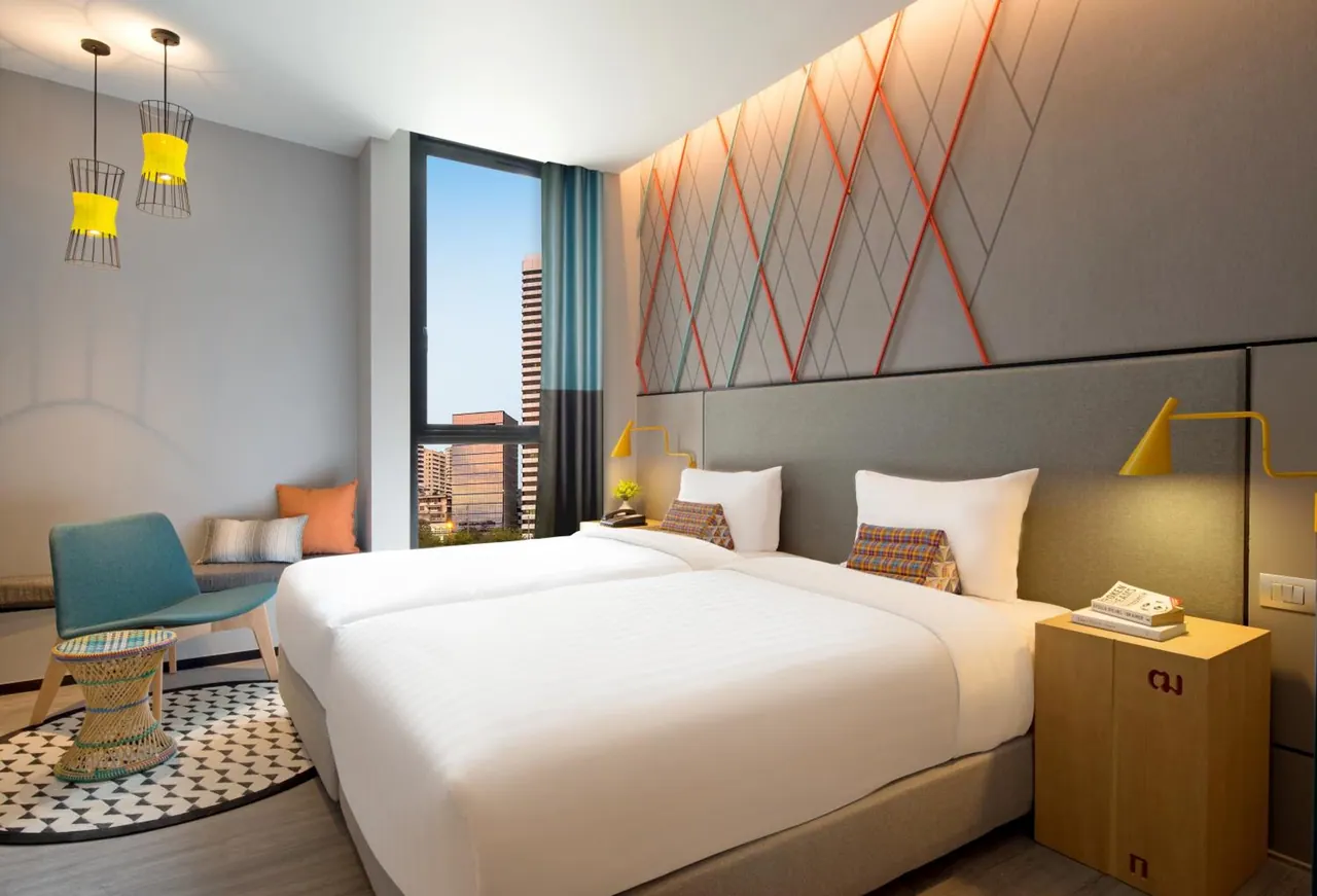 A bedroom with two single beds, a seating area by the window and a view of tall city buildings outside.