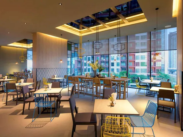 A dining room with square tables, coloured metal chairs, pendant lights and a skylight, with floor-to-ceiling windows looking onto residential buildings.