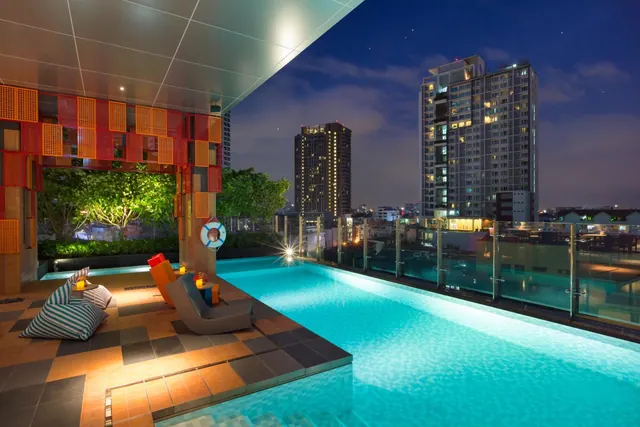 A rooftop swimming pool at night with lounge chairs, striped bean bags and city high-rise towers in the background.