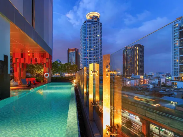 A long teal-tiled lap pool runs beside a glass railing overlooking illuminated city towers at dusk.