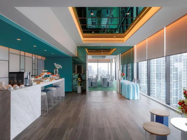 A lounge area with a marble counter, bar stools, a coffee machine and pastries on the counter, with high-rise buildings visible through the windows.