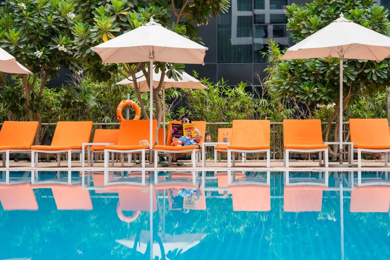 Orange sun loungers and cream parasols line the edge of a pool, with one guest reading on a lounger.