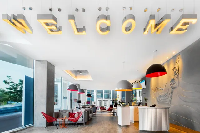 A lobby with illuminated WELCOME letters hanging from the ceiling, grey sofas, red chairs and a white curved reception desk.