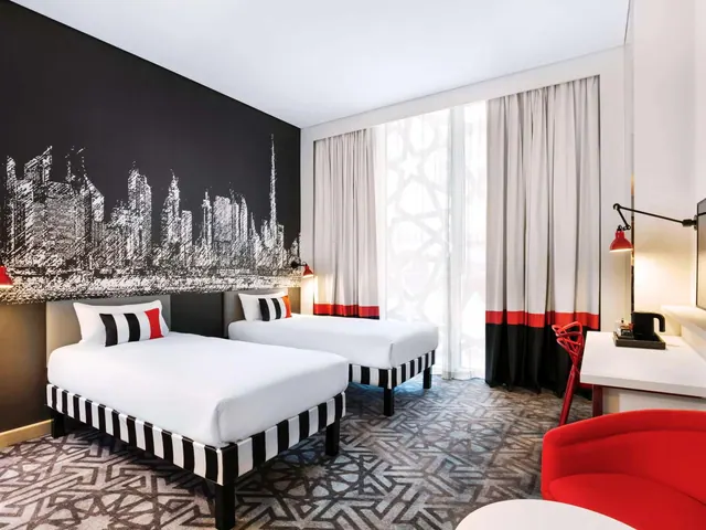 A hotel bedroom with two single beds with black and white striped bases, a city skyline mural on the wall, a red armchair, and a desk with a kettle.