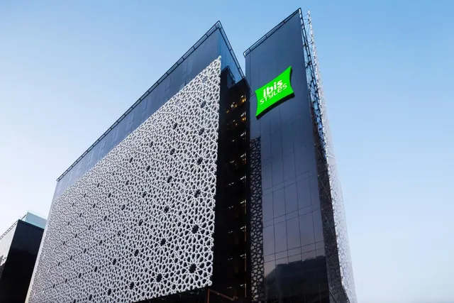 The exterior of an ibis Styles building with a white geometric lattice panel, dark glass facade and a green illuminated sign against a clear blue sky.
