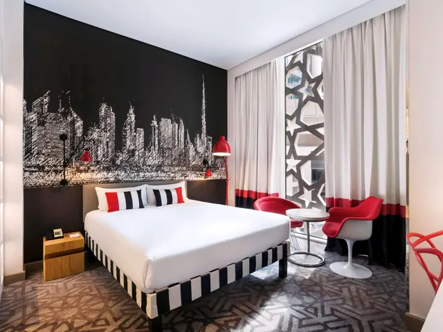 A bedroom with a made-up king bed, a black wall mural of a city skyline, red armchairs and a round table beside a tall window.