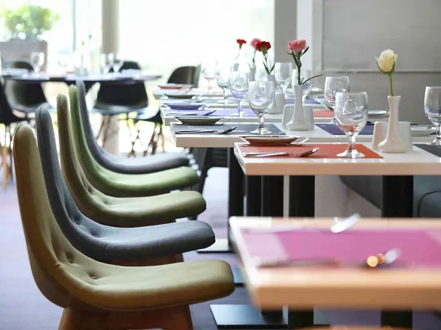 A restaurant dining room with set tables, wine glasses, small flower vases and rows of upholstered green and grey chairs.