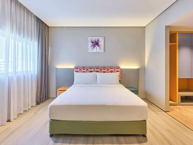 A made-up double bed with white linen sits against a grey wall, with sheer curtained windows on the left and an open wardrobe on the right.