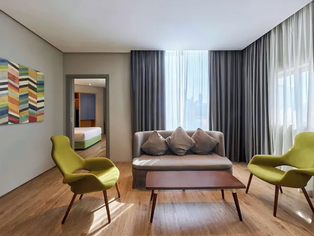 A living area with a grey sofa, two green armchairs and a wooden coffee table, with a bedroom visible through the doorway on the left.