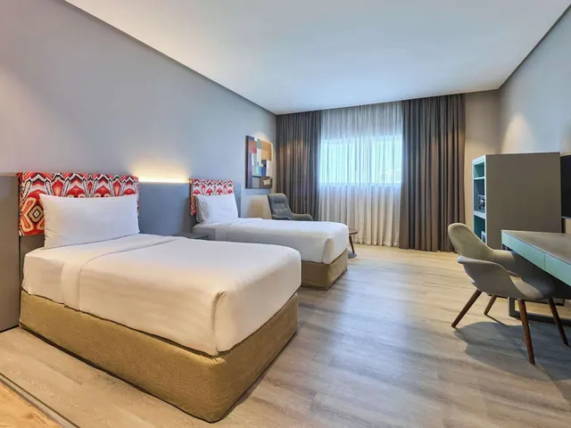 A hotel bedroom with two single beds with patterned red headboards, an armchair, a desk with two chairs and a storage shelf unit beside a window with sheer and dark curtains.
