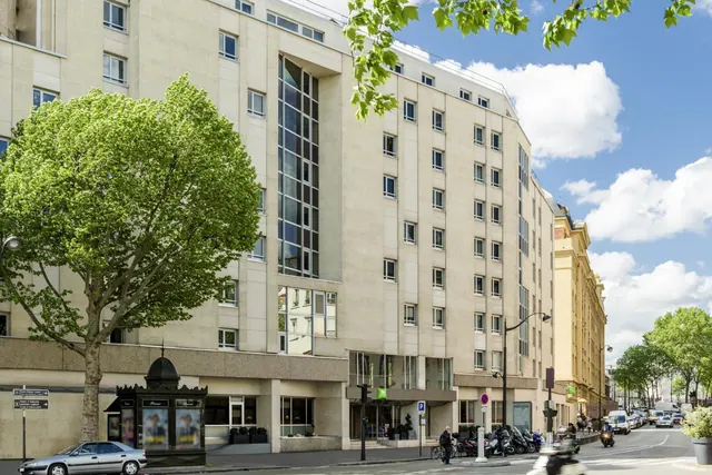A modern hotel building stands on a tree-lined Paris street beside older stone buildings.