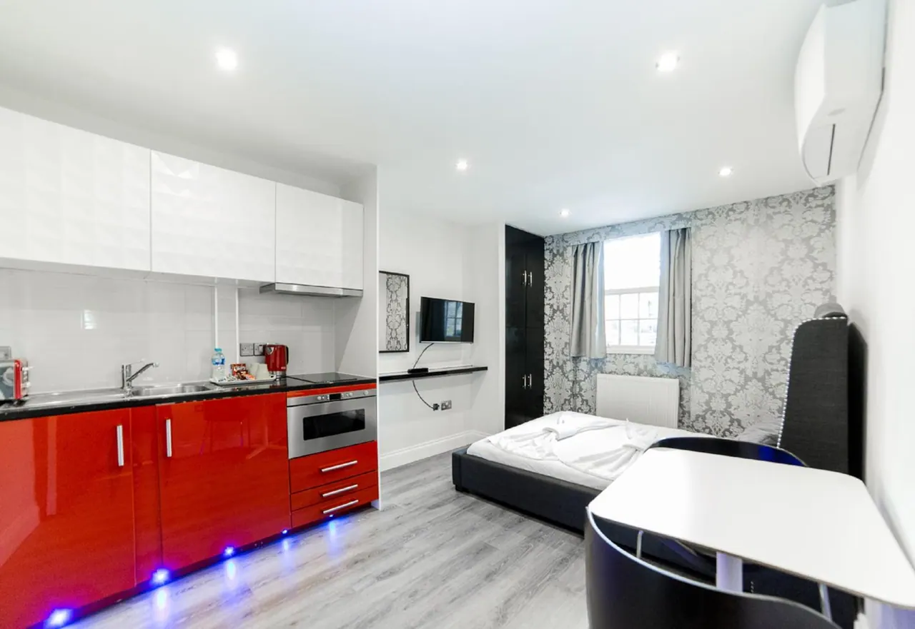 A studio room with a red and white kitchenette along one wall, a double bed with a black frame, a wall-mounted television, a desk with a chair, and patterned wallpaper around the window.