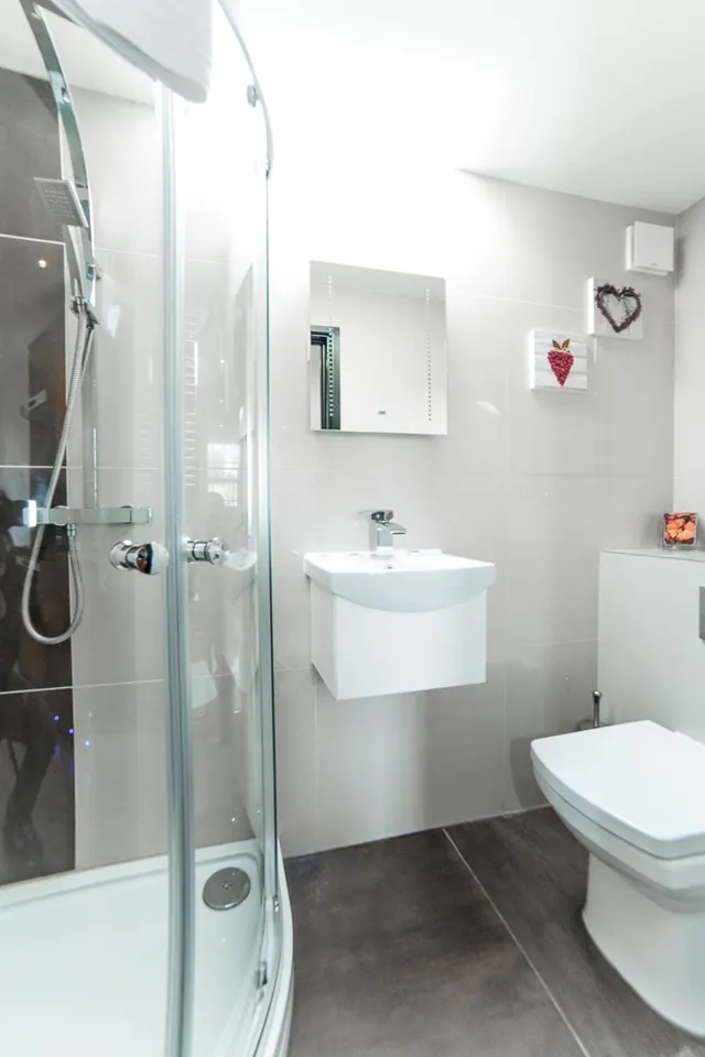 A bathroom with a curved glass shower enclosure, a wall-hung white sink with a mirror cabinet above, and a wall-mounted toilet with a small bidet spray beside it.