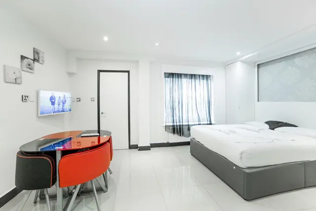 A white studio room with a double bed, a round glass dining table with red and dark chairs, a wall-mounted television and a window with sheer dark curtains.