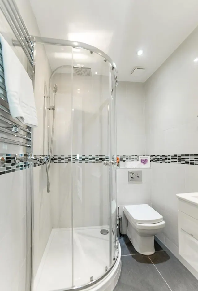 A bathroom with a curved glass corner shower enclosure, a wall-mounted toilet, a heated towel rail with white towels, and grey floor tiles.