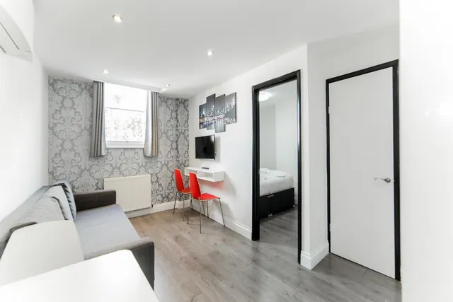 A living area with a grey sofa, a wall-mounted television, a small white wall desk with two red chairs, and an open doorway to a bedroom with a bed.