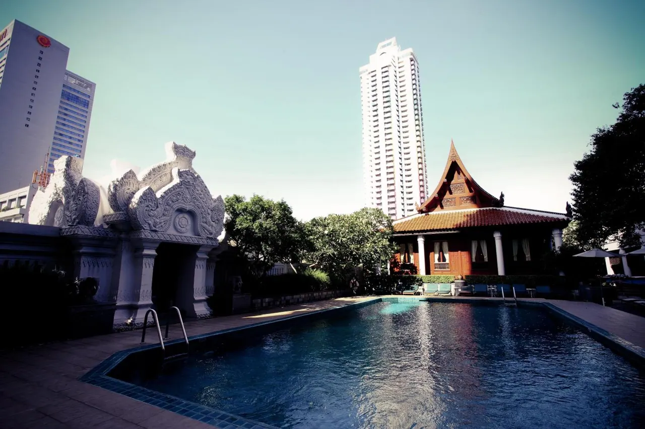 A rectangular outdoor swimming pool sits beside a Thai-style pavilion and an ornate white gateway, with a tall tower rising behind them.