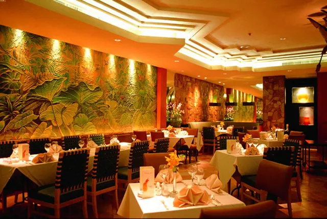 A restaurant dining room with a large tropical leaf mural on one wall, with set tables, folded napkins and wine glasses.