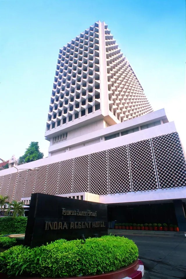 The exterior of the Indra Regent Hotel tower, with a patterned concrete facade, seen from a low angle under a clear blue sky, with the hotel name on a dark sign in front of shrubbery.