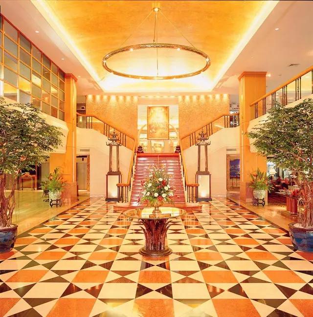 A hotel lobby with a checkered marble floor, a central staircase, a large ring chandelier, potted trees and a flower arrangement on a round table.