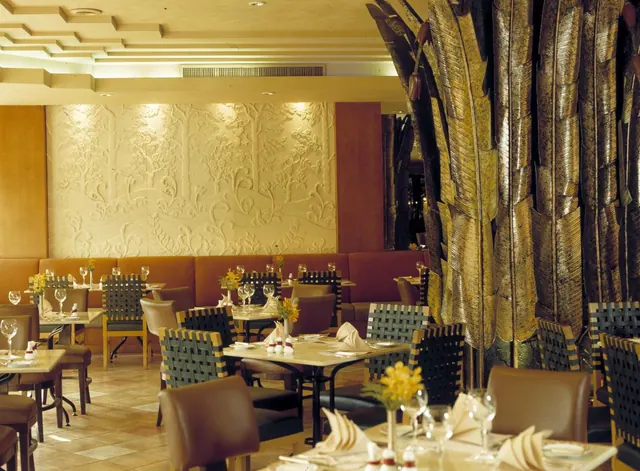 A restaurant dining room with set tables, woven-back chairs, a leather banquette along the wall, a carved relief wall and tall metallic leaf sculptures.