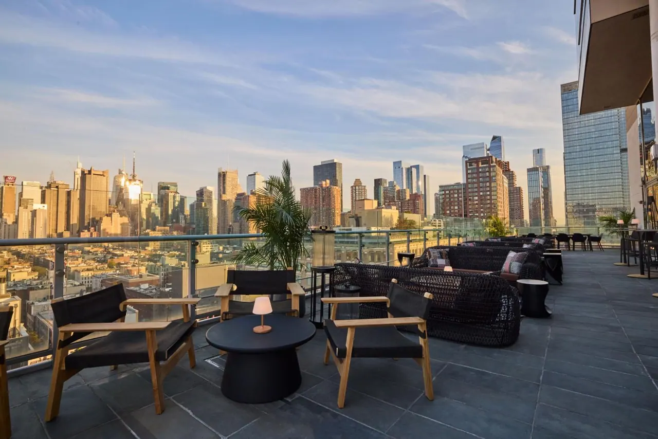A rooftop terrace with wooden and black woven lounge chairs, low tables and a view of a city skyline in warm evening light.