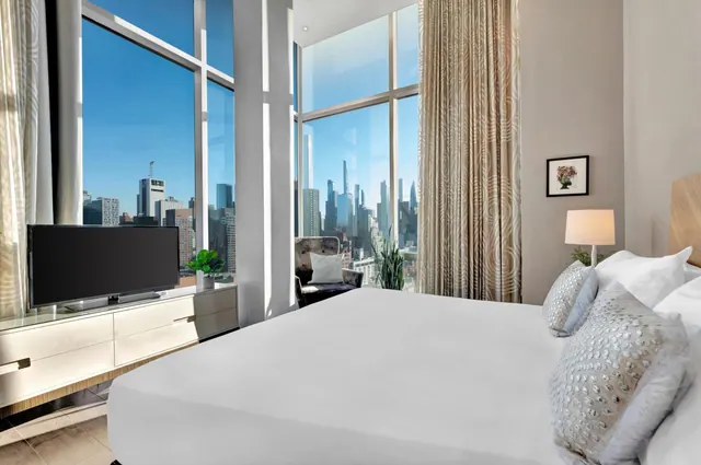 A bedroom with a made king bed, a flat-screen television on a low sideboard, and floor-to-ceiling windows showing a city skyline.