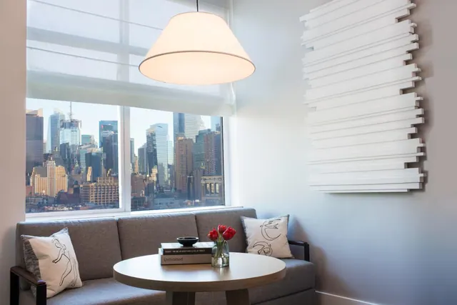 A seating area with a grey sofa, a round wooden table holding books and a vase of red tulips, and a window showing a city skyline.