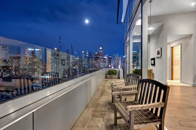 A terrace with wooden benches and a glass railing overlooks a lit city skyline under a night sky with a moon.