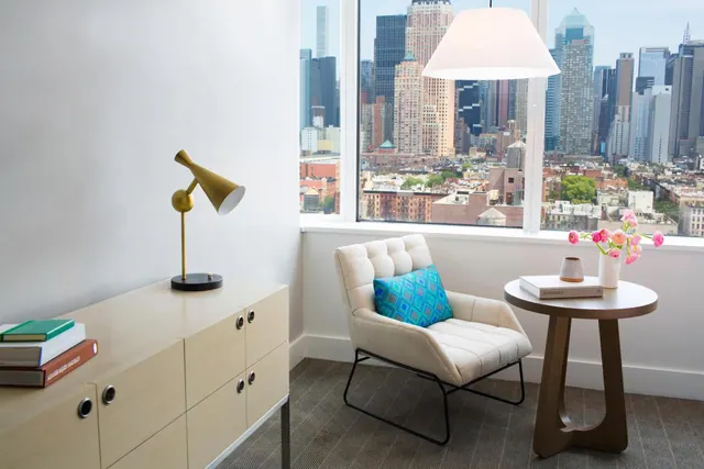 A sitting area with a cream armchair, a blue patterned cushion, a round side table with flowers, and a brass table lamp, beside a window showing a city skyline.