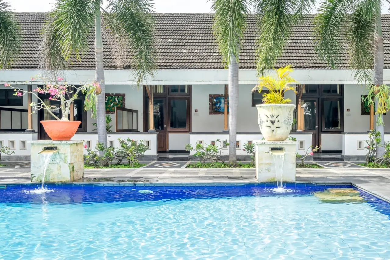 A blue-tiled swimming pool sits in front of a single-storey white building with a tiled roof, with two stone water spouts, potted plants and palm trees along the walkway.