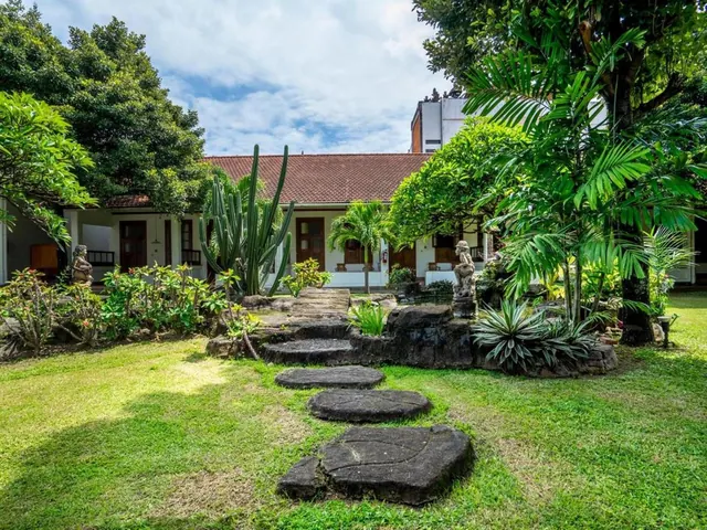 A stone stepping-stone path crosses a lawn toward a low white building with a red tile roof, flanked by cacti, palms and garden statues.